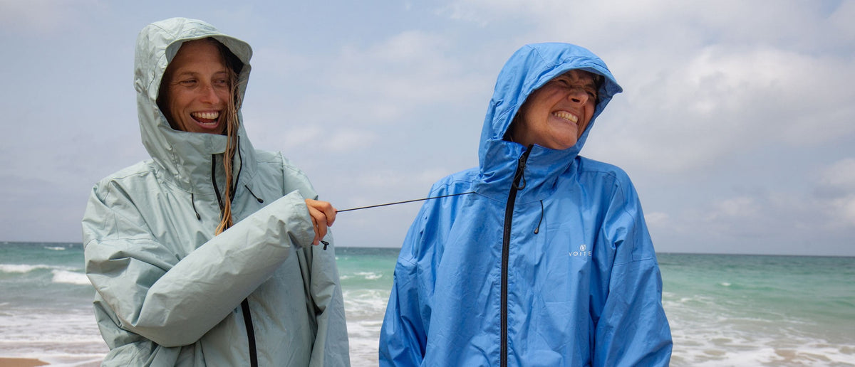 Zwei Personen tragen leichte Regenjacken mit Kapuze der Marke Allweather Coat und stehen lachend am Strand, während eine Person an der Zugschnur der Jacke der anderen zieht. Die Jacke links ist blassmintgrün, die Jacke rechts mittelblau.