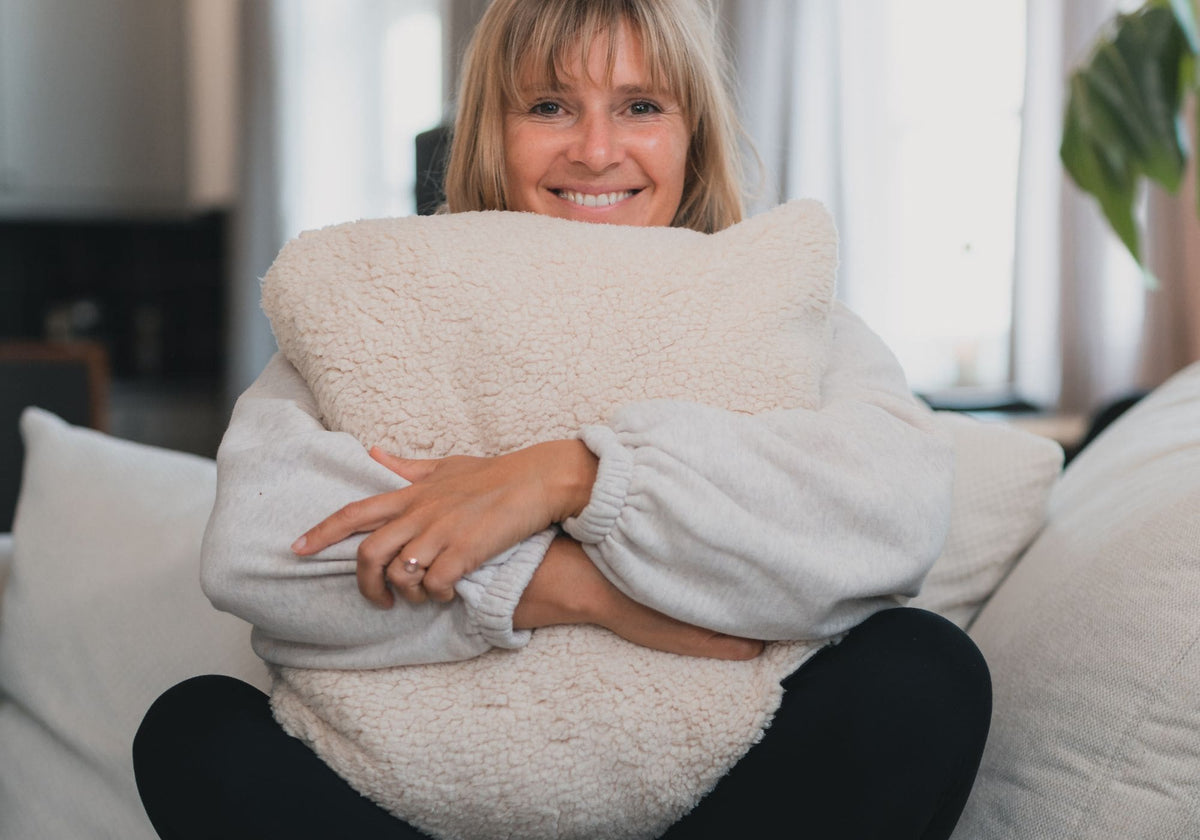 Lächelnde Frau in hellgrauem Strickpullover umarmt großes cremefarbenes VOITED Cloudtouch Sherpa-Kissen auf Sofa.