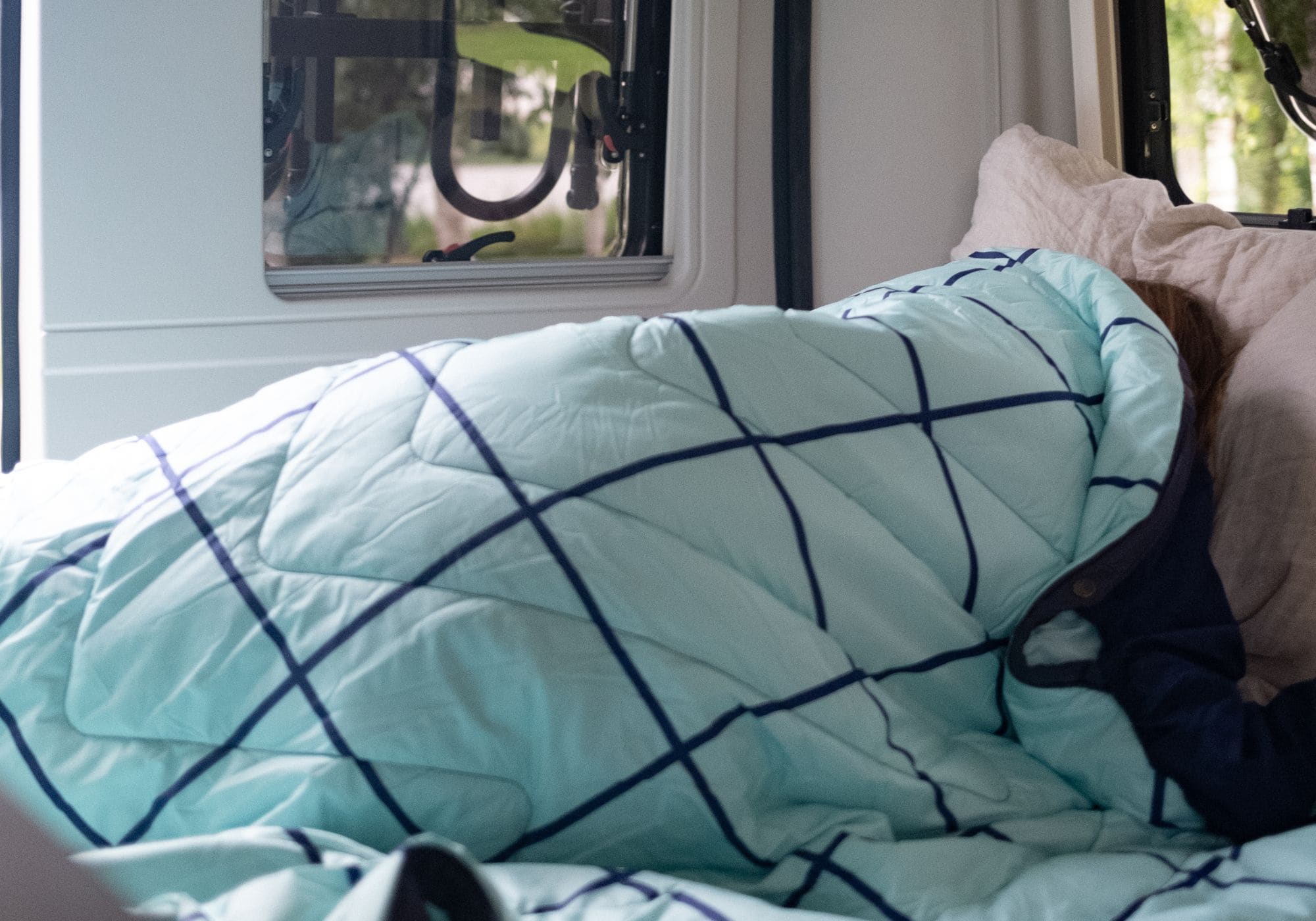 Hellblaue gesteppte Bettdecke mit dunkelblauem Gittermuster QUEEN Cloudtouch VOITED POOL auf einem Bett, darunter teilweise eine Person zu sehen, daneben ein beiges Kissen