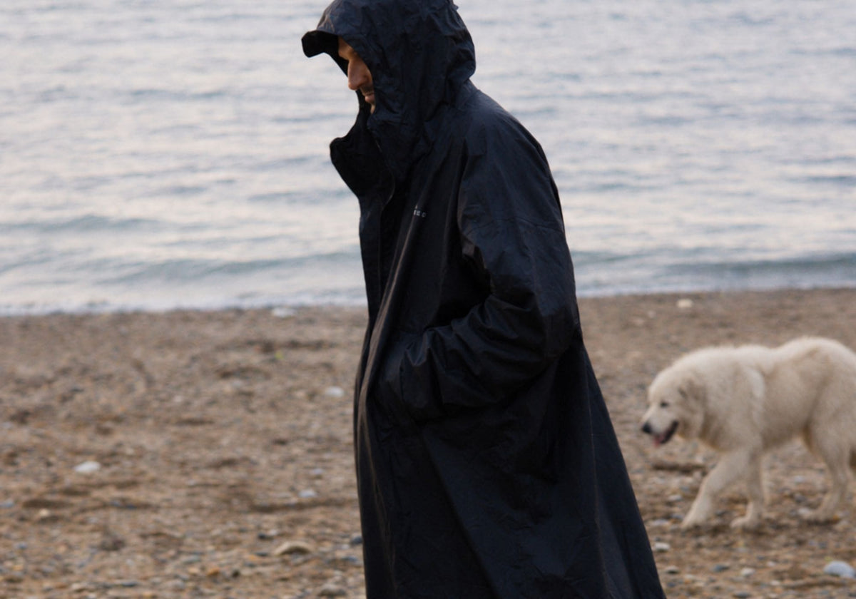 Mann im schwarzen VOITED Allweather Coat beim Spaziergang am Strand, daneben ein weißer Hund. AI modified