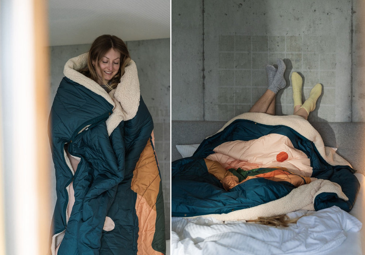 Frau eingehüllt in eine VOITED Cloudtouch Decke mit kuscheligem Sherpa-Futter; zwei Personen liegen unter der Decke im Bett, die Füße in Socken an einer Betonwand.
