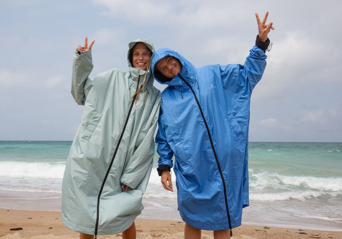 Zwei lachende Frauen in VOITED Allweather Coats in Mintgrün und Blau am Strand, mit Peace-Zeichen vor dem Meer
