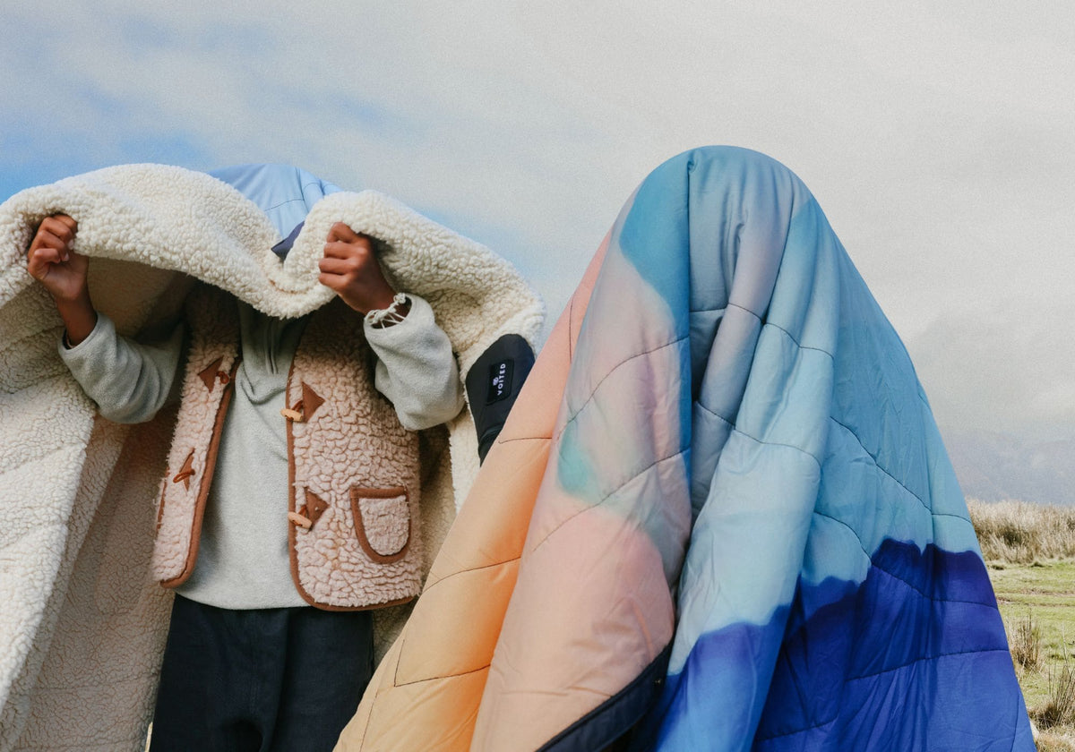 Zwei Personen draußen unter bewölktem Himmel. Eine hüllt sich in eine flauschige Teddyfleece-Decke, die andere ist komplett unter einer gesteppten Voited Cloudtouch Decke mit Farbverlauf von Pfirsich über Hellblau zu Dunkelblau verborgen.