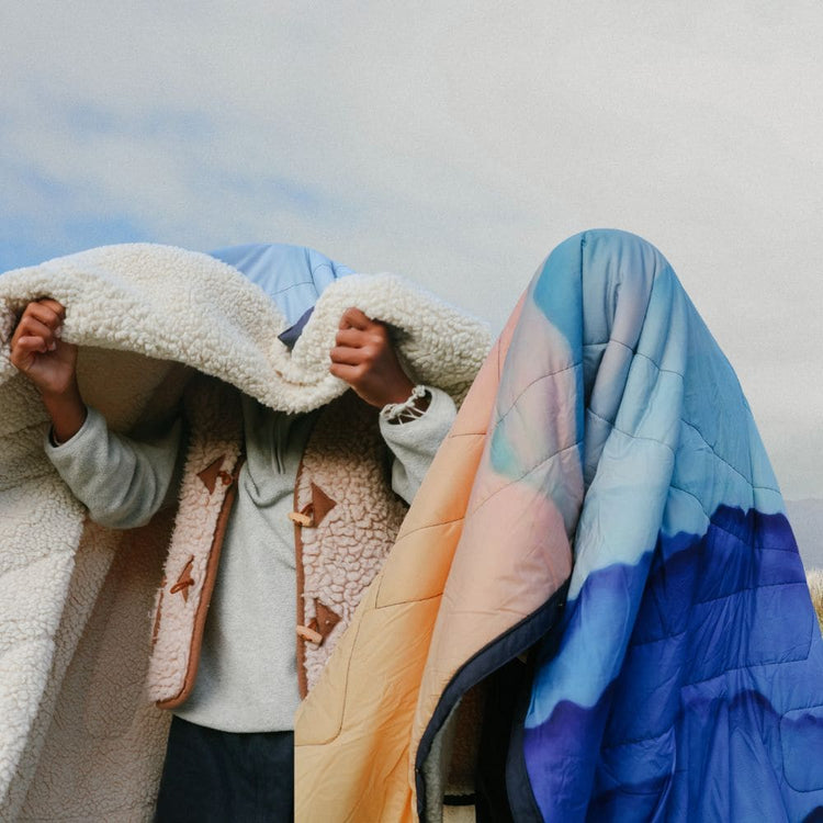 Zwei Personen draußen unter bewölktem Himmel. Eine hüllt sich in eine flauschige Teddyfleece-Decke, die andere ist komplett unter einer gesteppten Voited Cloudtouch Decke mit Farbverlauf von Pfirsich über Hellblau zu Dunkelblau verborgen.