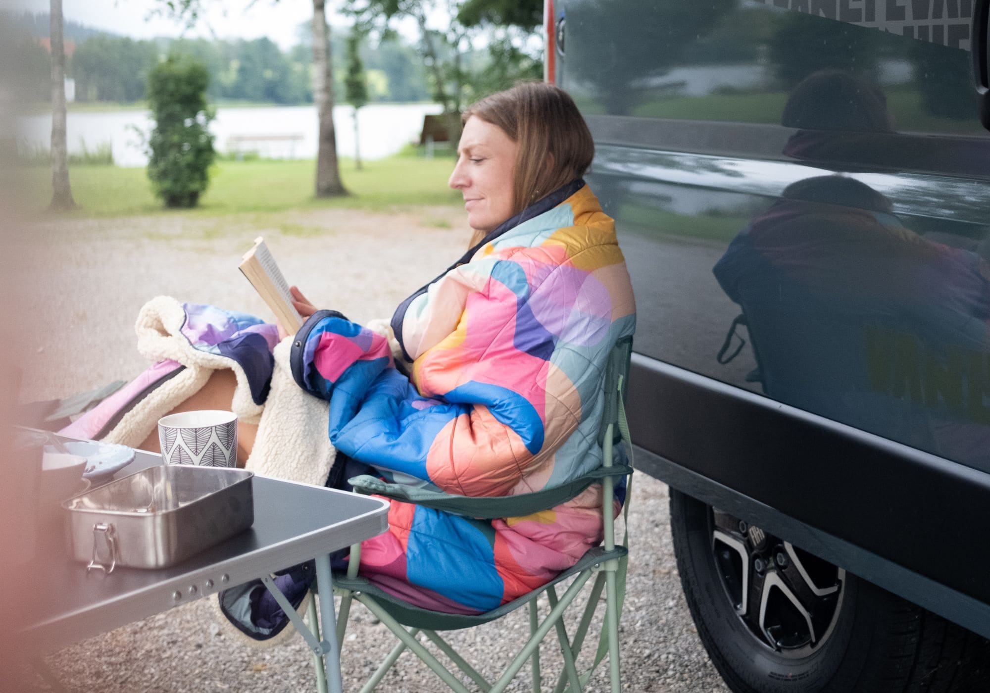 Frau in bunter gesteppter VOITED Cloudtouch Decke liest in Campingstuhl neben Fahrzeug, im Hintergrund Bäume und Wasser.