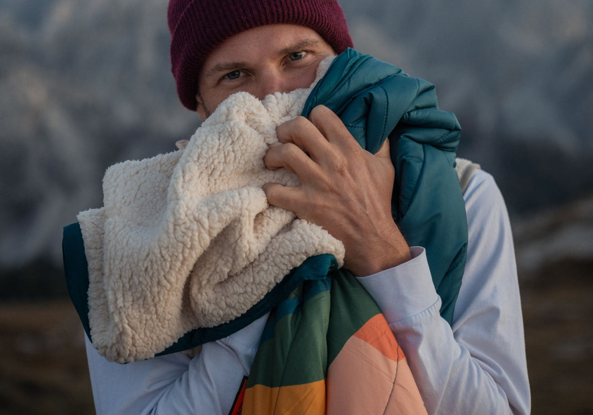 Mann mit Mütze hält eine VOITED Cloudtouch Decke mit cremefarbener Sherpa- und türkis-oranger Seite, Berge im Hintergrund.