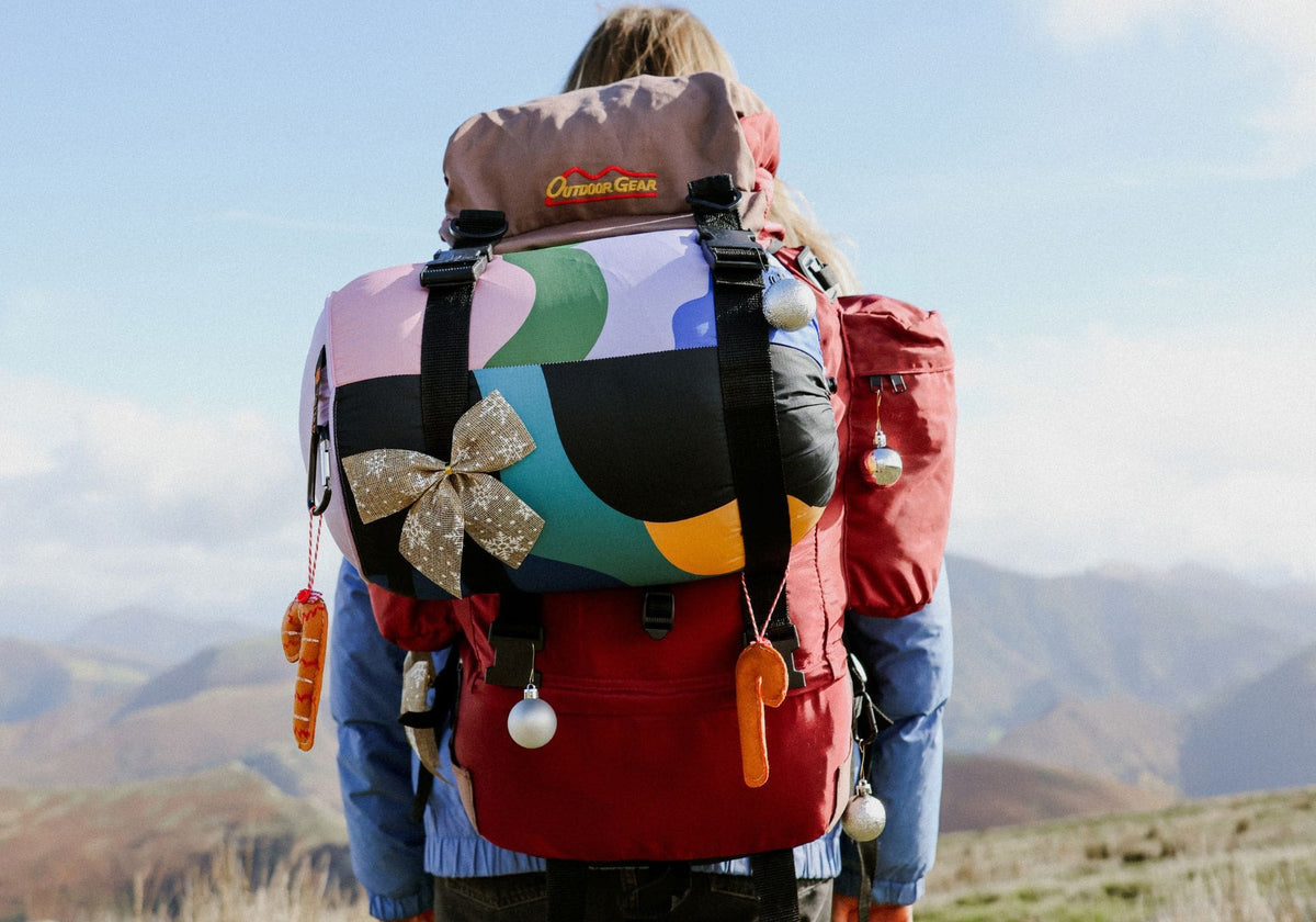 Wanderin in den Bergen mit bunter VOITED Cloudtouch Decke auf einem weihnachtlich geschmückten Rucksack.