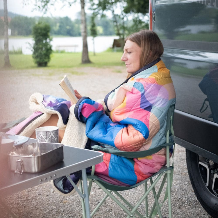 Frau in bunter gesteppter VOITED Cloudtouch Decke liest in Campingstuhl neben Fahrzeug, im Hintergrund Bäume und Wasser.