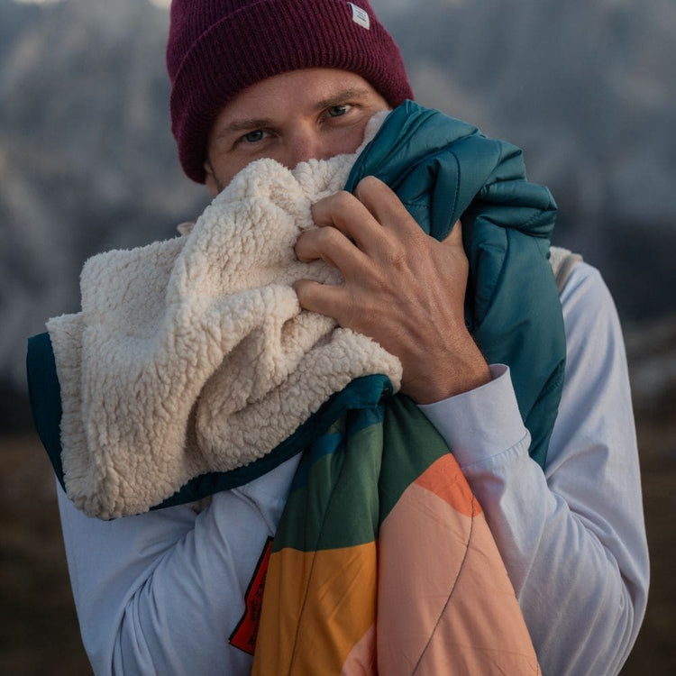 Mann mit Mütze hält eine VOITED Cloudtouch Decke mit cremefarbener Sherpa- und türkis-oranger Seite, Berge im Hintergrund.