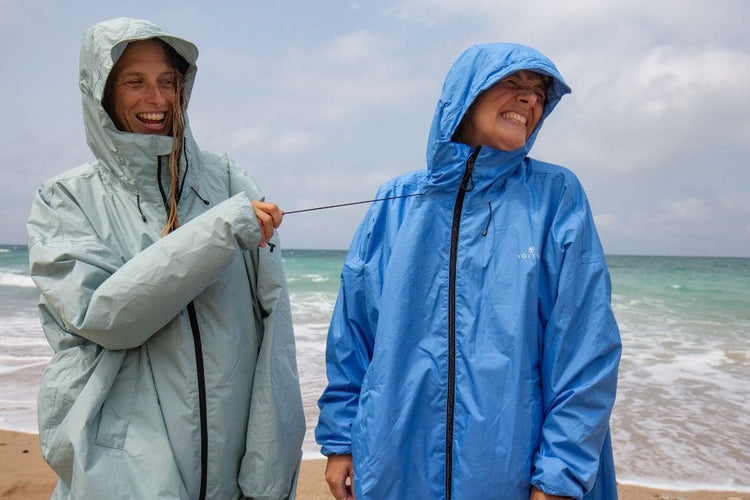 Zwei Personen tragen leichte Regenjacken mit Kapuze der Marke Allweather Coat und stehen lachend am Strand, während eine Person an der Zugschnur der Jacke der anderen zieht. Die Jacke links ist blassmintgrün, die Jacke rechts mittelblau.
