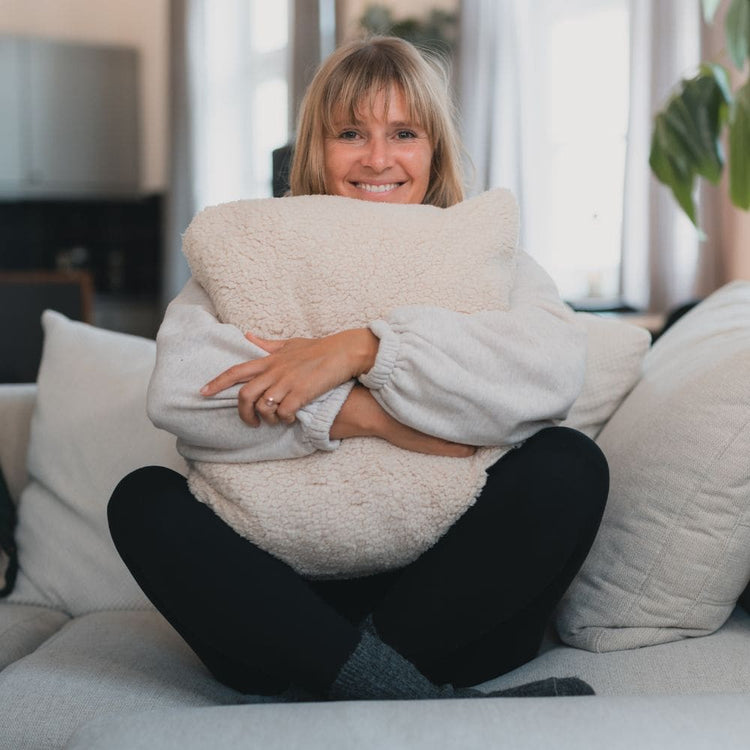 Lächelnde Frau in hellgrauem Strickpullover umarmt großes cremefarbenes VOITED Cloudtouch Sherpa-Kissen auf Sofa.