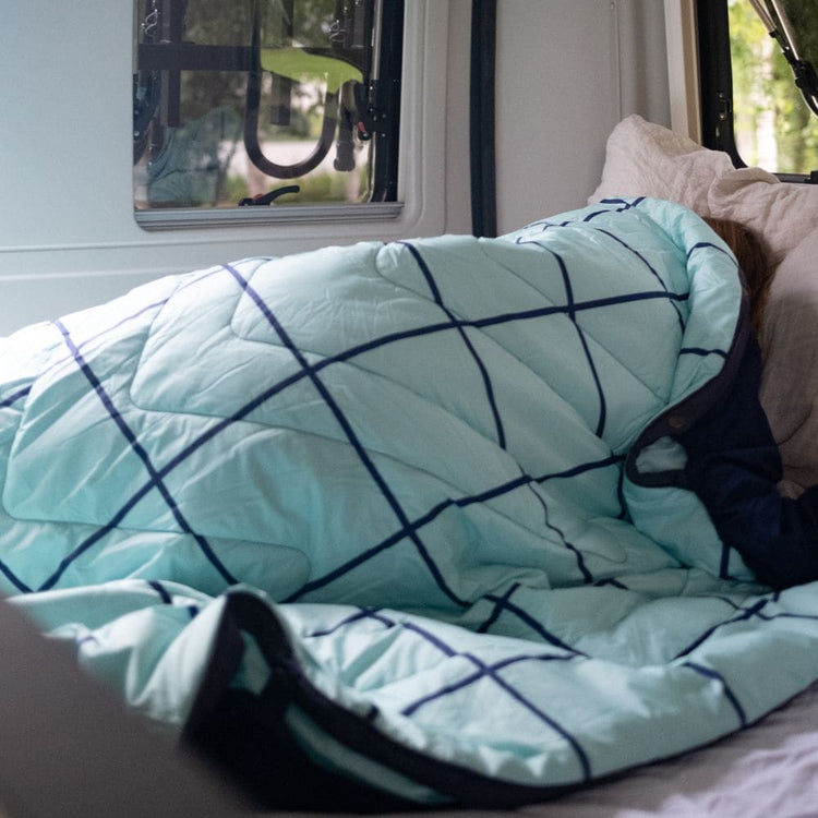 Hellblaue gesteppte Bettdecke mit dunkelblauem Gittermuster QUEEN Cloudtouch VOITED POOL auf einem Bett, darunter teilweise eine Person zu sehen, daneben ein beiges Kissen