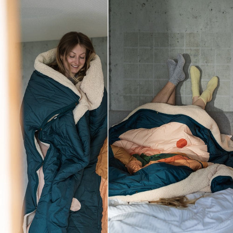 Frau eingehüllt in eine VOITED Cloudtouch Decke mit kuscheligem Sherpa-Futter; zwei Personen liegen unter der Decke im Bett, die Füße in Socken an einer Betonwand.