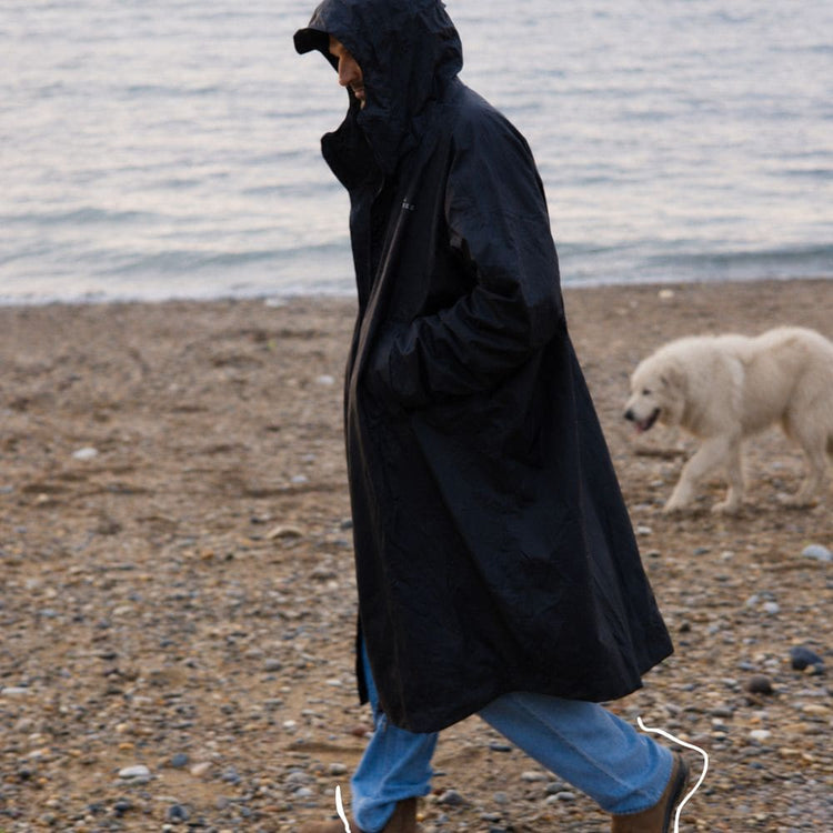 Mann im schwarzen VOITED Allweather Coat beim Spaziergang am Strand, daneben ein weißer Hund. AI modified