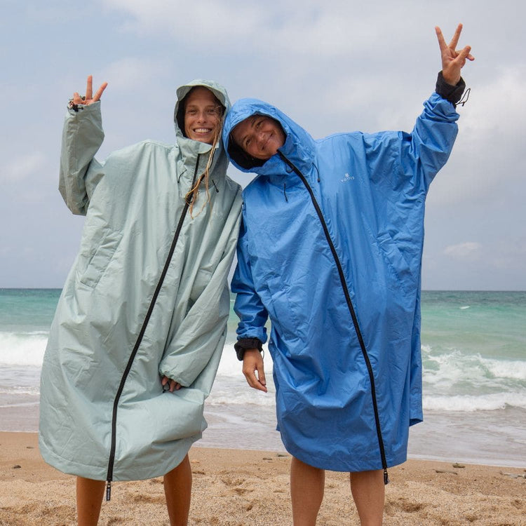 Zwei lachende Frauen in VOITED Allweather Coats in Mintgrün und Blau am Strand, mit Peace-Zeichen vor dem Meer
